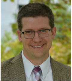 Man with brown hair wearing a plaid suit, white dress shirt, and purple tie.