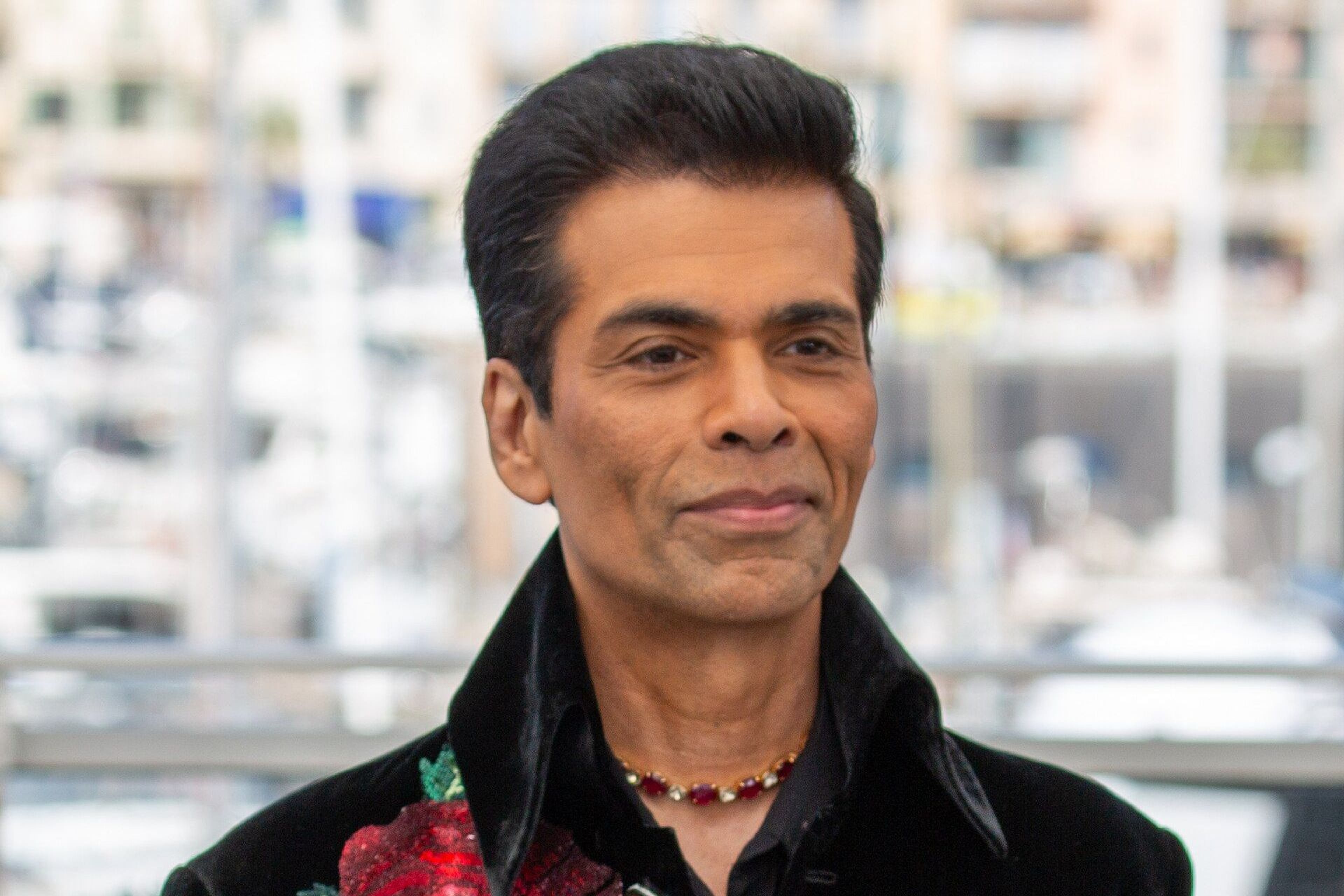 Karan Johar in a black velvet jacket with red rose embroidery, standing before a blurred city waterfront backdrop.