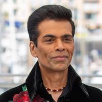 Karan Johar in a black velvet jacket with red rose embroidery, standing before a blurred city waterfront backdrop.