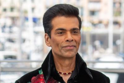 Karan Johar in a black velvet jacket with red rose embroidery, standing before a blurred city waterfront backdrop.