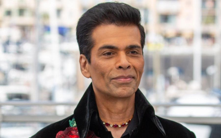 Karan Johar in a black velvet jacket with red rose embroidery, standing before a blurred city waterfront backdrop.