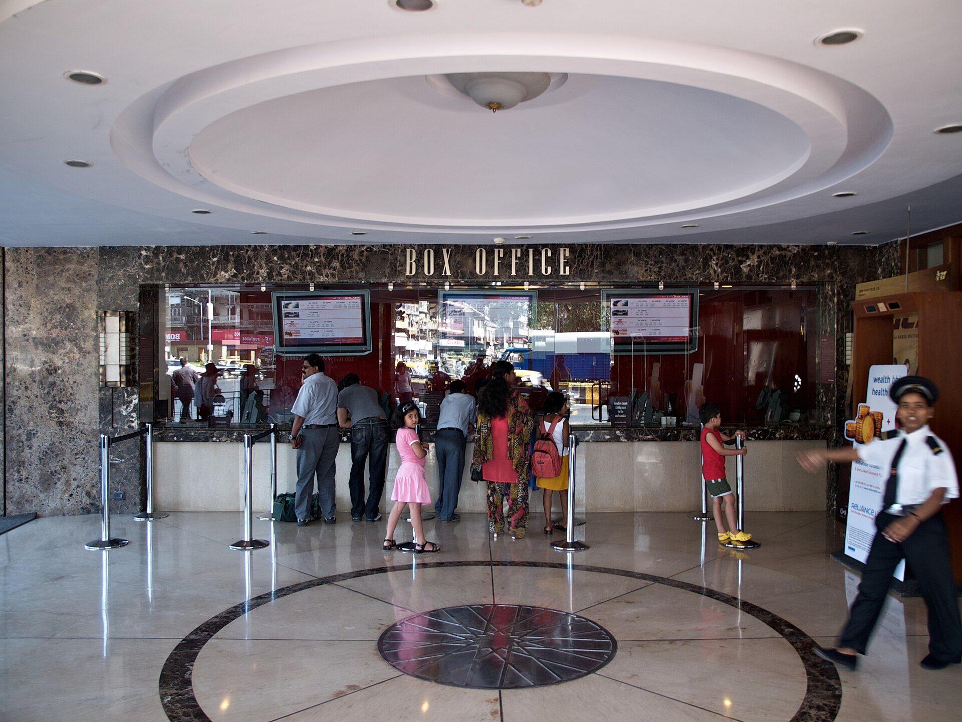 People stand at a cinema box office counter in a marble-floored lobby while a uniformed guard walks past on the right