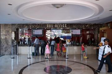 People stand at a cinema box office counter in a marble-floored lobby while a uniformed guard walks past on the right