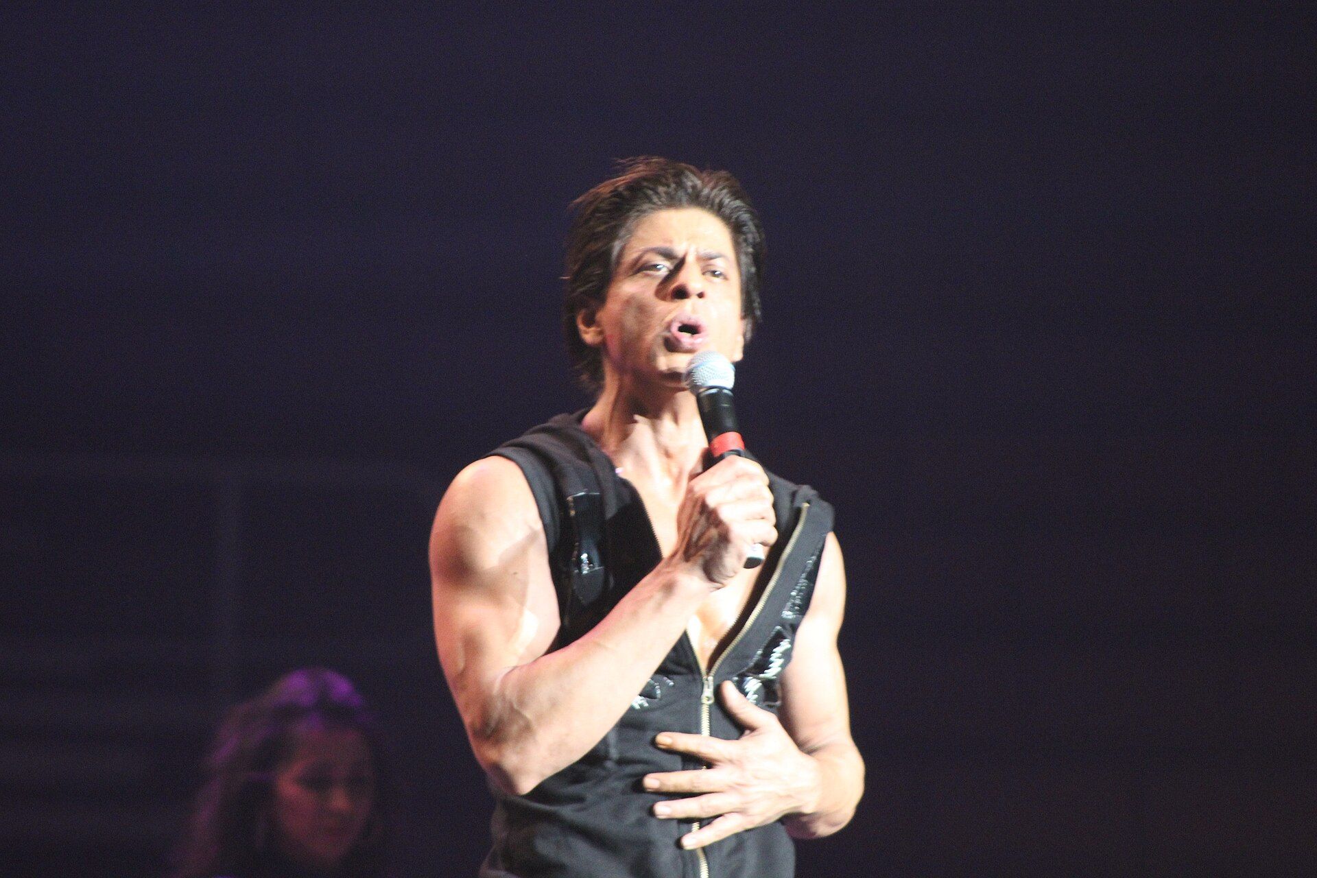 Shah Rukh Khan sings into a microphone on a dark stage, in a sleeveless black top