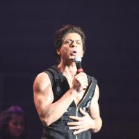 Shah Rukh Khan sings into a microphone on a dark stage, in a sleeveless black top