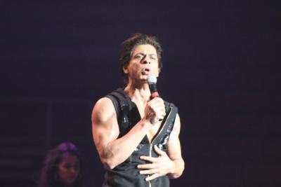 Shah Rukh Khan sings into a microphone on a dark stage, in a sleeveless black top