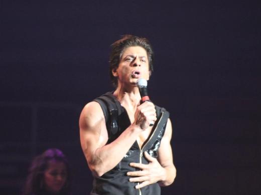 Shah Rukh Khan sings into a microphone on a dark stage, in a sleeveless black top