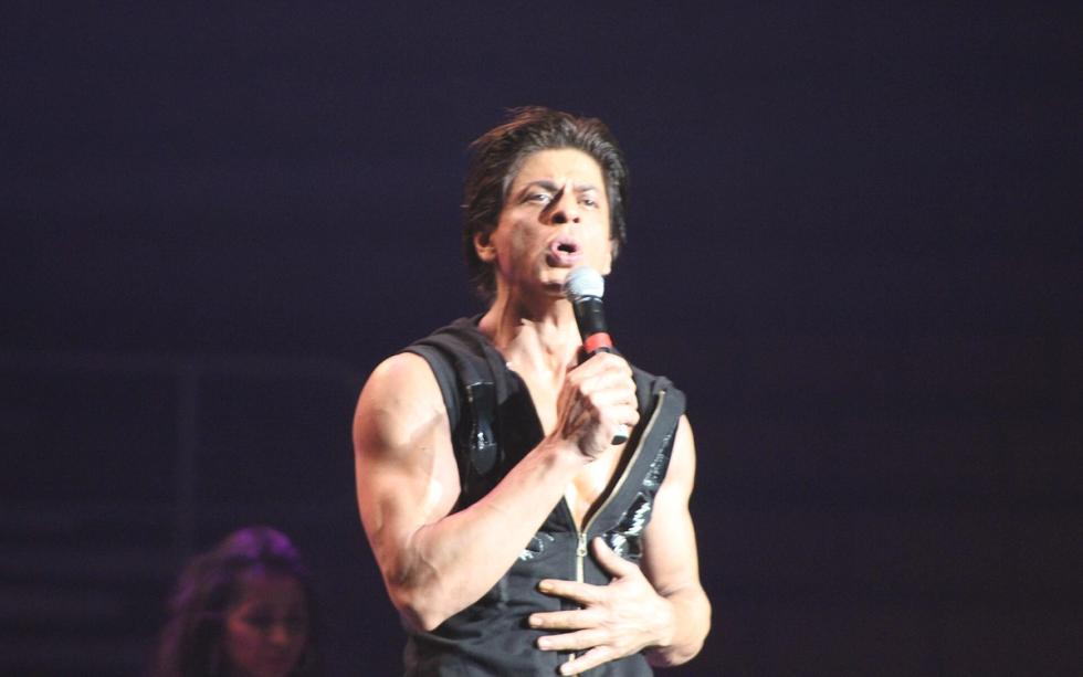 Shah Rukh Khan sings into a microphone on a dark stage, in a sleeveless black top