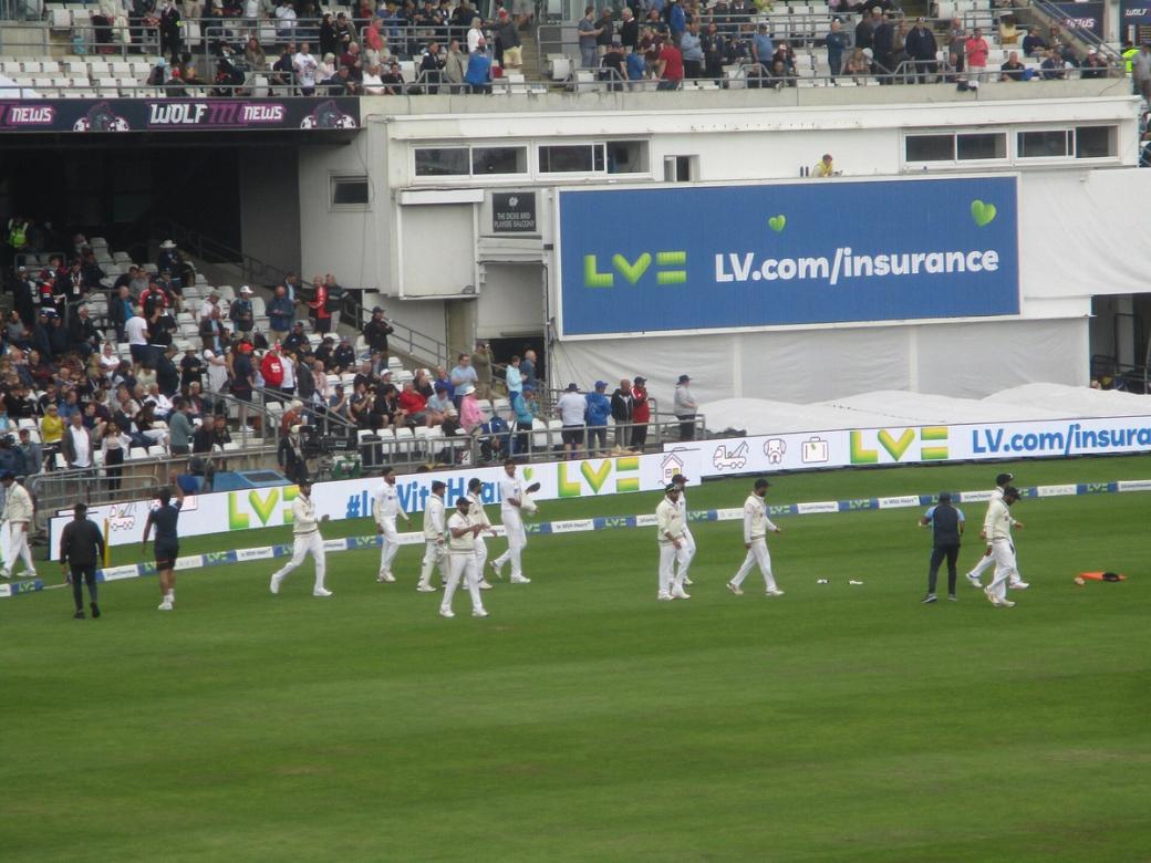 Indian players in white kit walk across the field during a Test match, with spectators in the stands.