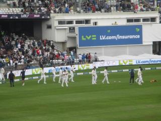 Indian players in white kit walk across the field during a Test match, with spectators in the stands.