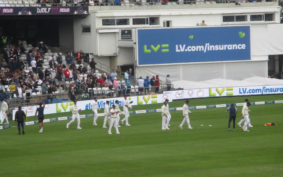 Indian players in white kit walk across the field during a Test match, with spectators in the stands.