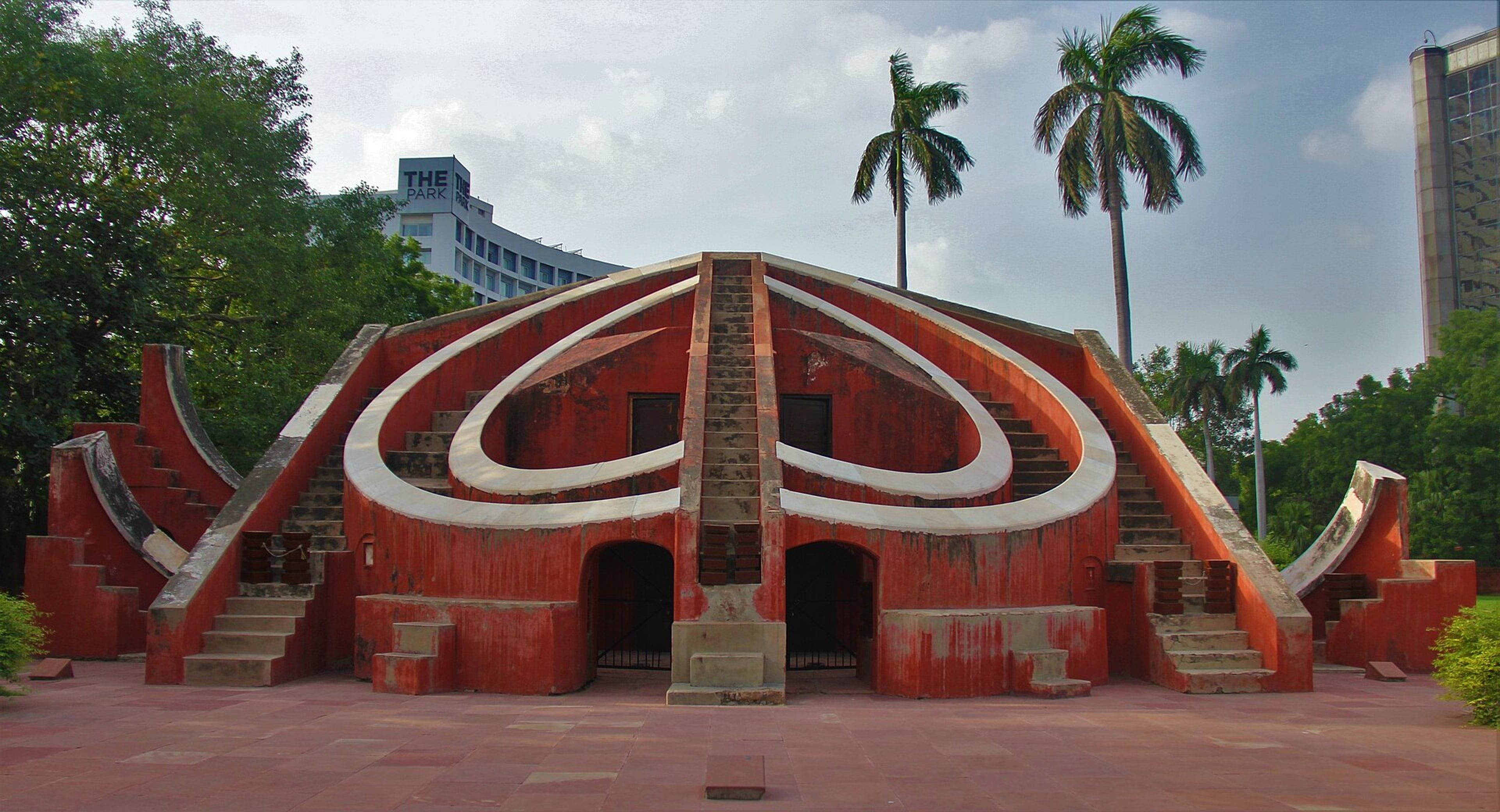 Red sandstone Misra Yantra stone observatory with curved walls and stairs, palm trees and a hotel tower behind.