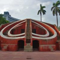 Red sandstone Misra Yantra stone observatory with curved walls and stairs, palm trees and a hotel tower behind.