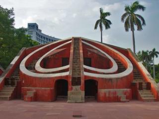 Red sandstone Misra Yantra stone observatory with curved walls and stairs, palm trees and a hotel tower behind.