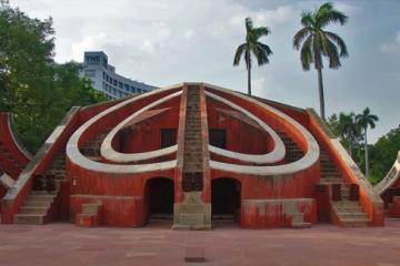 Red sandstone Misra Yantra stone observatory with curved walls and stairs, palm trees and a hotel tower behind.