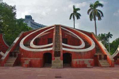 Red sandstone Misra Yantra stone observatory with curved walls and stairs, palm trees and a hotel tower behind.