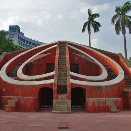 Red sandstone Misra Yantra stone observatory with curved walls and stairs, palm trees and a hotel tower behind.