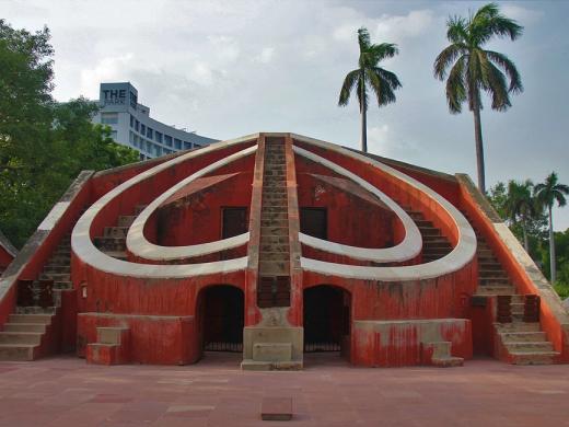 Red sandstone Misra Yantra stone observatory with curved walls and stairs, palm trees and a hotel tower behind.