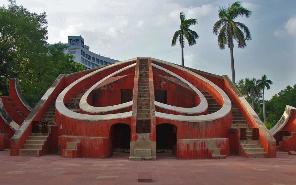 Red sandstone Misra Yantra stone observatory with curved walls and stairs, palm trees and a hotel tower behind.