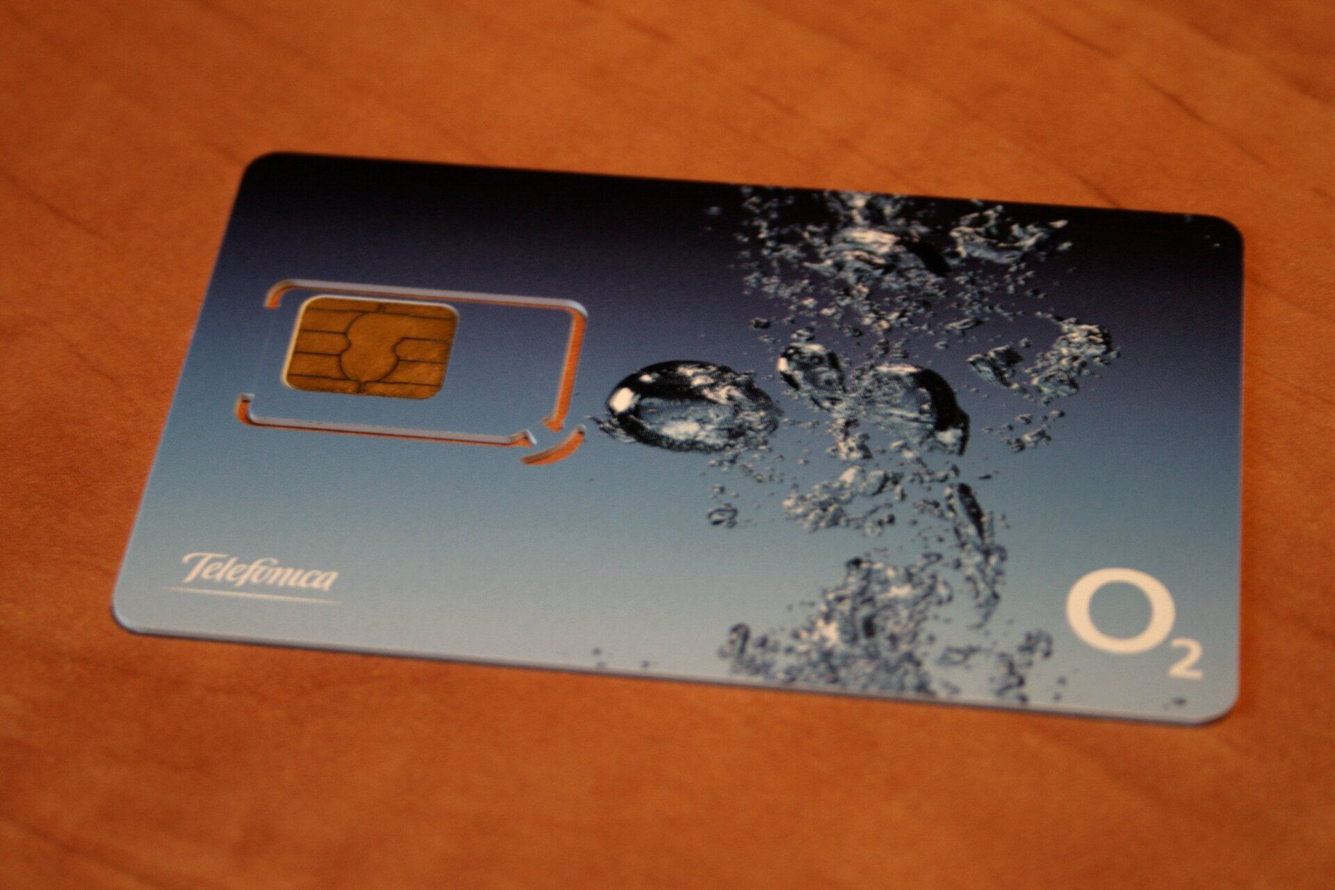 A blue prepaid SIM card with water droplets and a Telefónica O2 logo, lying on a wooden table