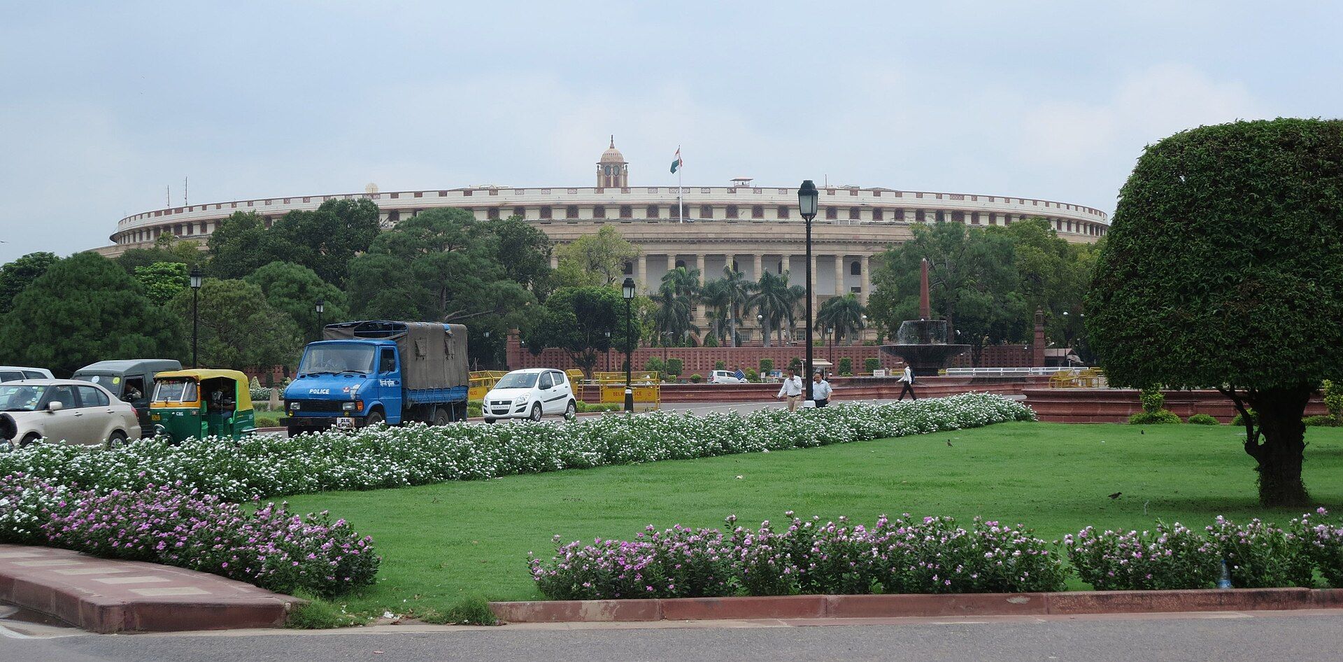 Curved colonnaded Parliament building in New Delhi with a flag on its dome, lawns, flower beds and traffic in front