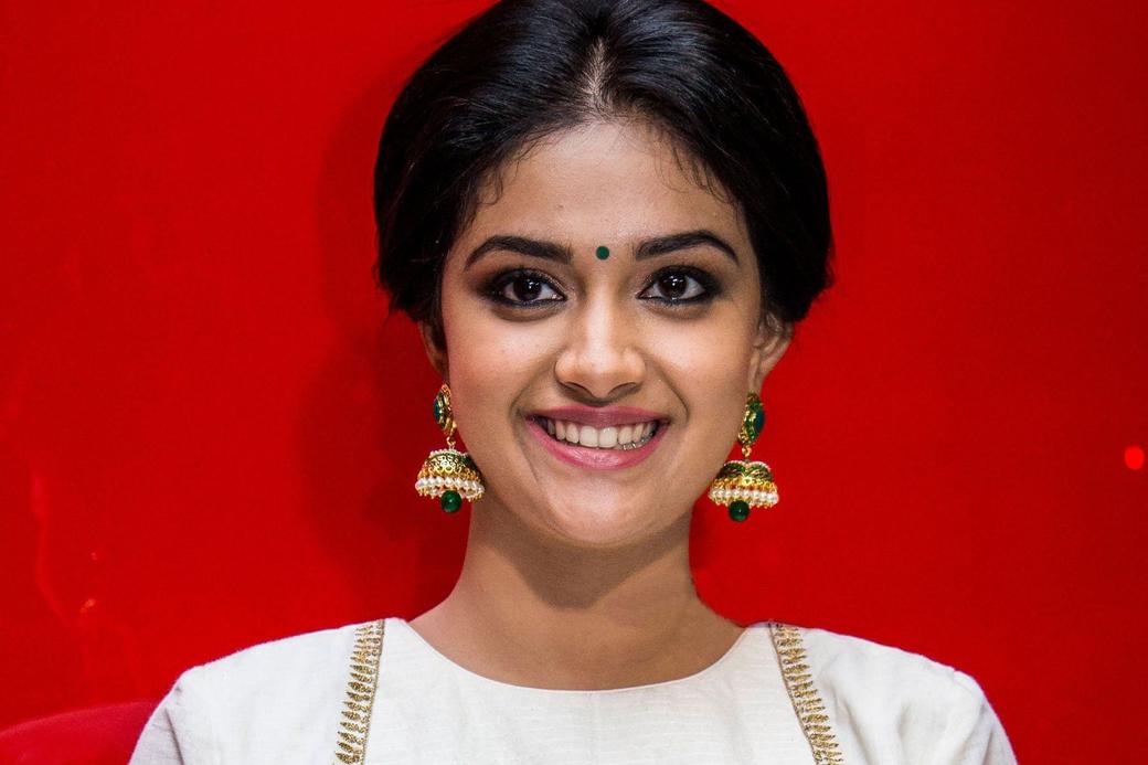 Keerthy Suresh smiles in a white and gold blouse and cream skirt against a bright red backdrop.