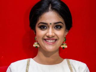 Keerthy Suresh smiles in a white and gold blouse and cream skirt against a bright red backdrop.