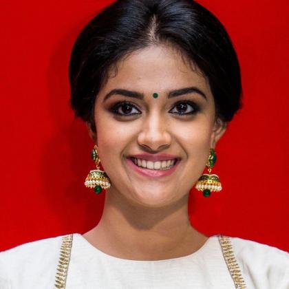 Keerthy Suresh smiles in a white and gold blouse and cream skirt against a bright red backdrop.