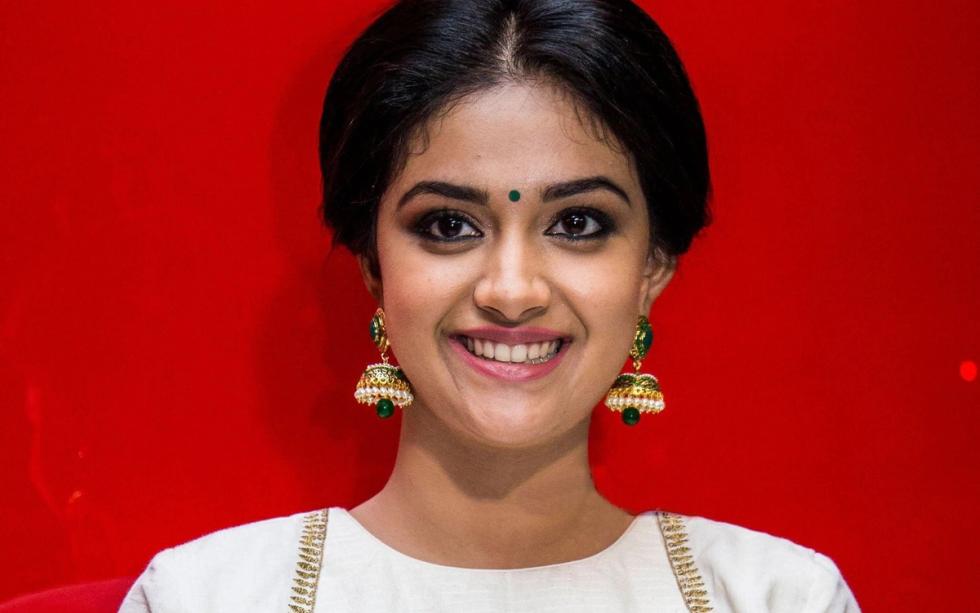 Keerthy Suresh smiles in a white and gold blouse and cream skirt against a bright red backdrop.