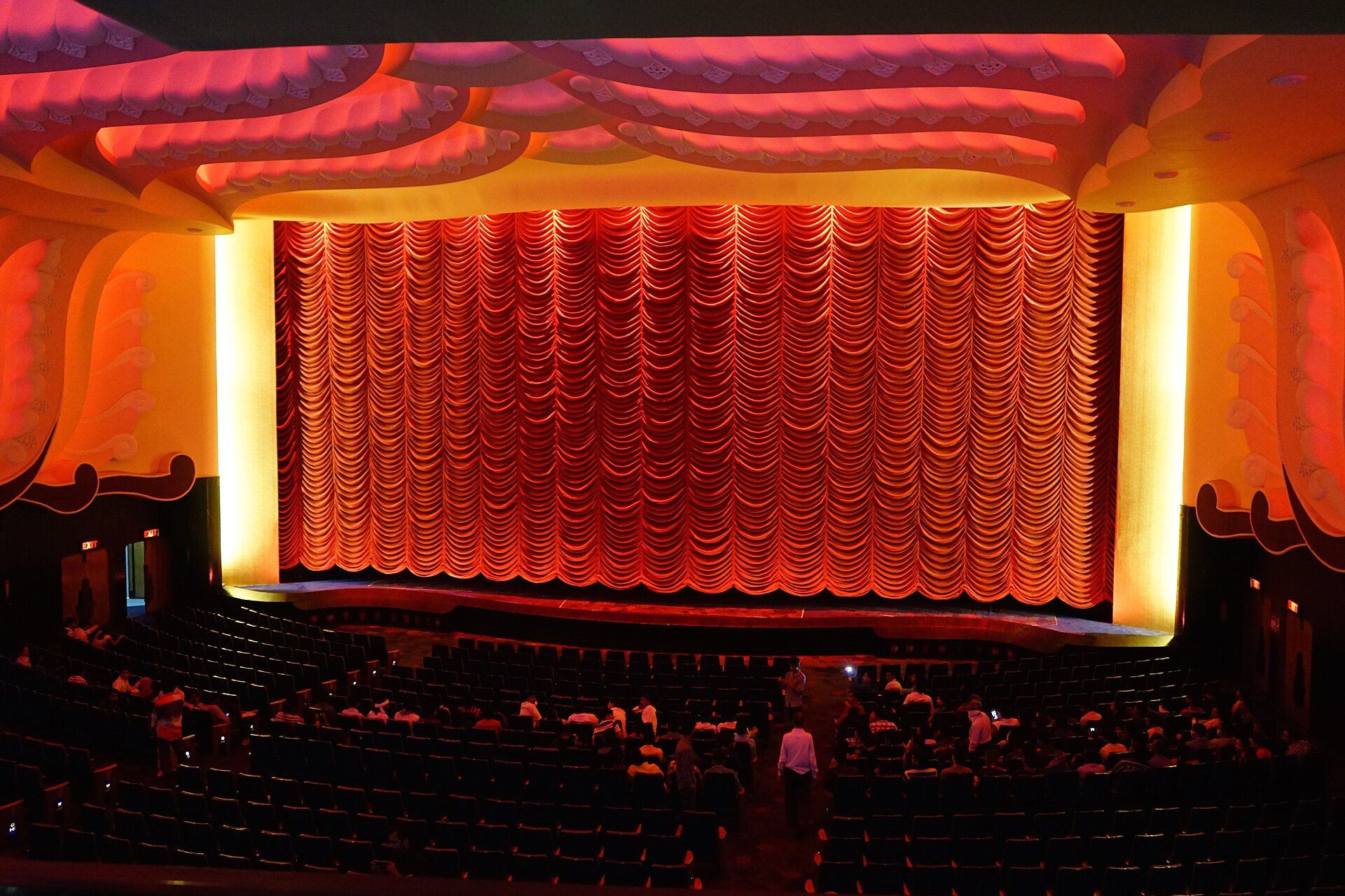 Rows of seats facing a lit red curtain across the screen inside an ornate cinema auditorium