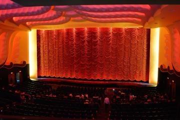 Rows of seats facing a lit red curtain across the screen inside an ornate cinema auditorium