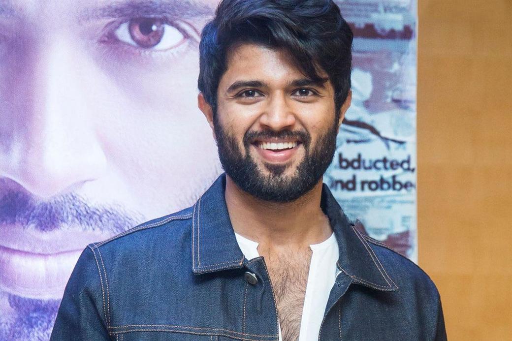 Smiling man in a denim jacket and white shirt stands in front of a film poster at a press meet.