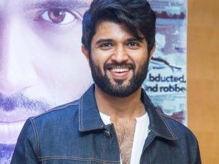 Smiling man in a denim jacket and white shirt stands in front of a film poster at a press meet.