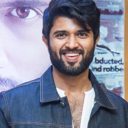 Smiling man in a denim jacket and white shirt stands in front of a film poster at a press meet.