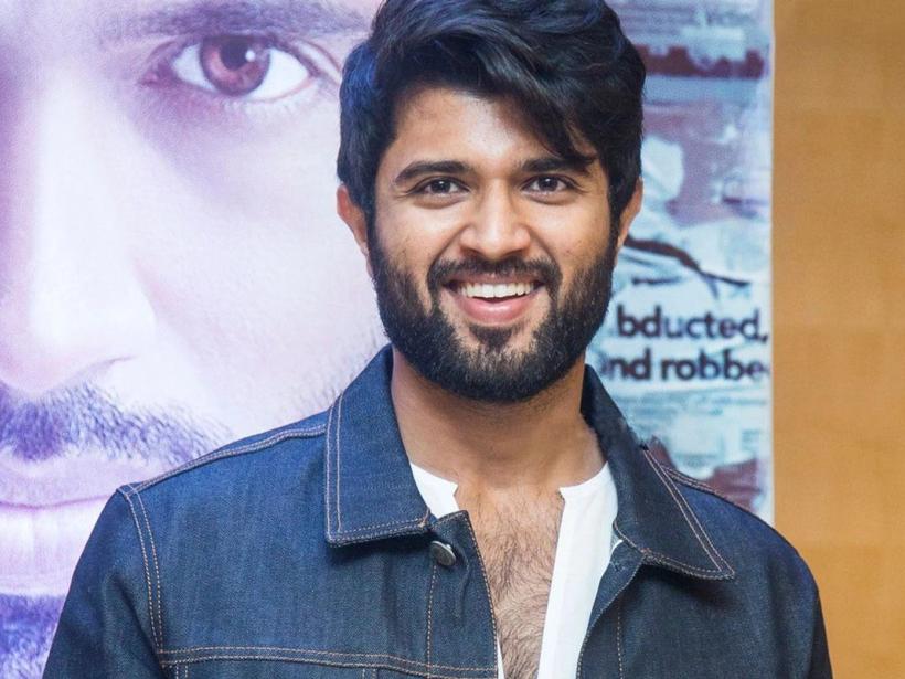 Smiling man in a denim jacket and white shirt stands in front of a film poster at a press meet.