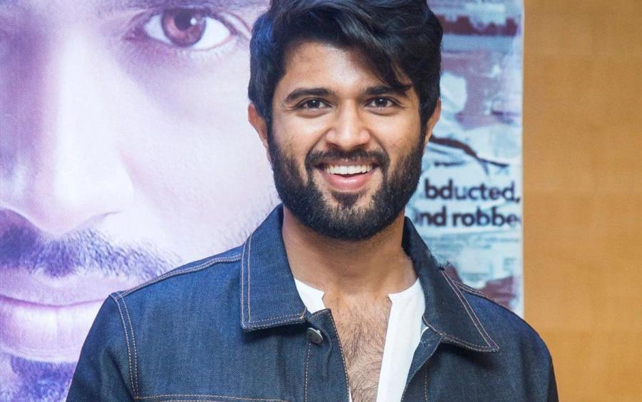 Smiling man in a denim jacket and white shirt stands in front of a film poster at a press meet.