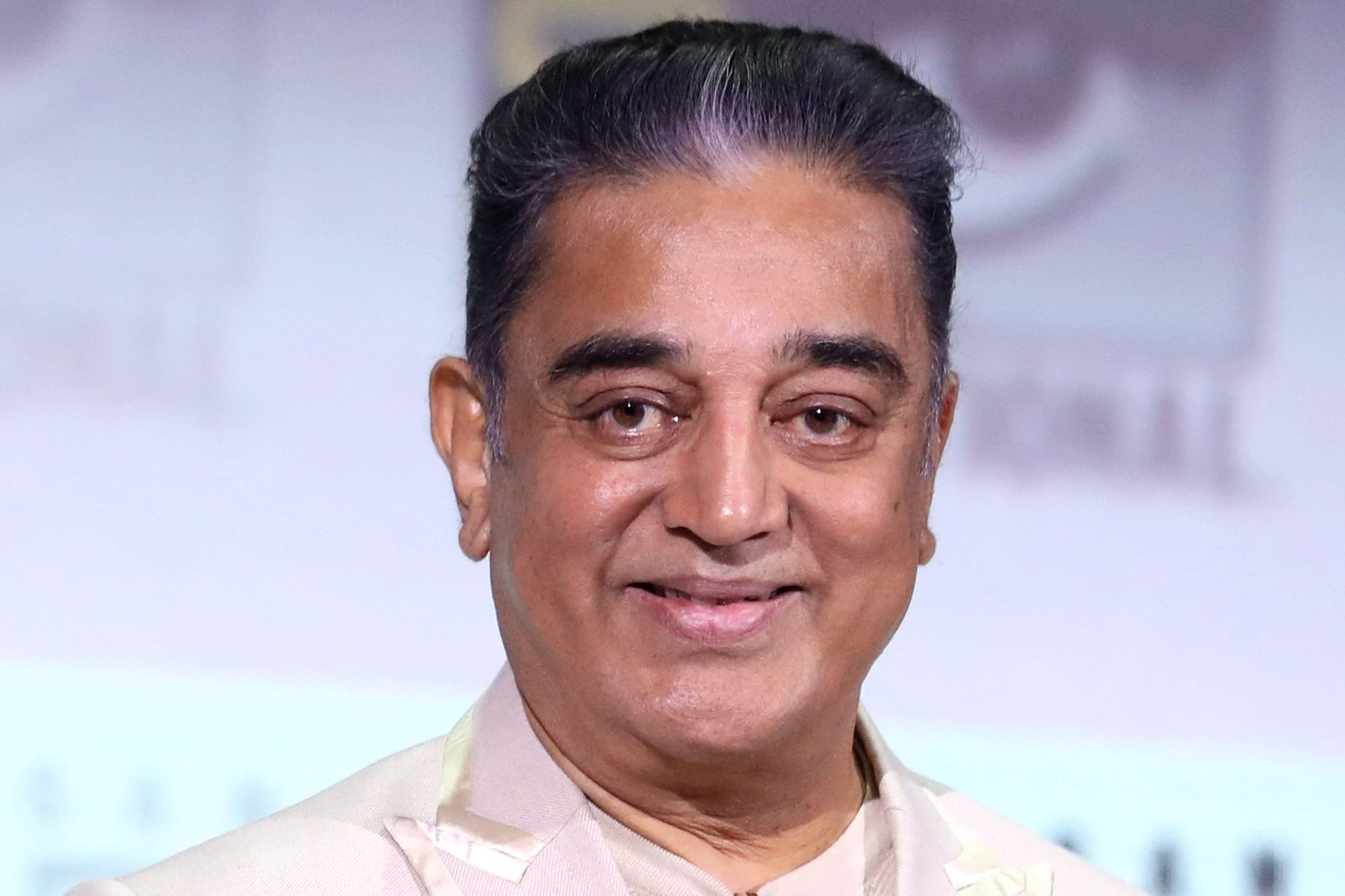 Kamal Haasan in a cream blazer with sunglasses hanging from his collar, smiling against a light backdrop.