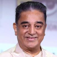 Kamal Haasan in a cream blazer with sunglasses hanging from his collar, smiling against a light backdrop.