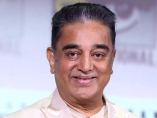 Kamal Haasan in a cream blazer with sunglasses hanging from his collar, smiling against a light backdrop.