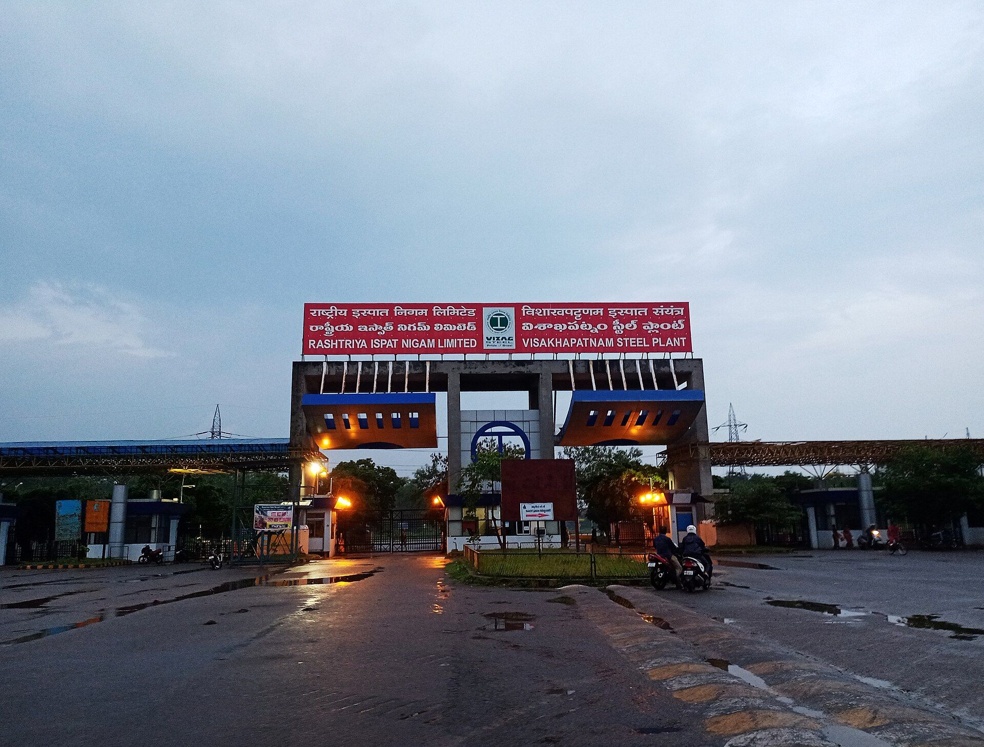 Entrance gate of the Visakhapatnam Steel Plant at dusk, with a red signboard, wet road and two people on a motorcycle