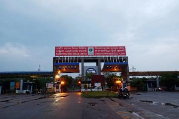 Entrance gate of the Visakhapatnam Steel Plant at dusk, with a red signboard, wet road and two people on a motorcycle