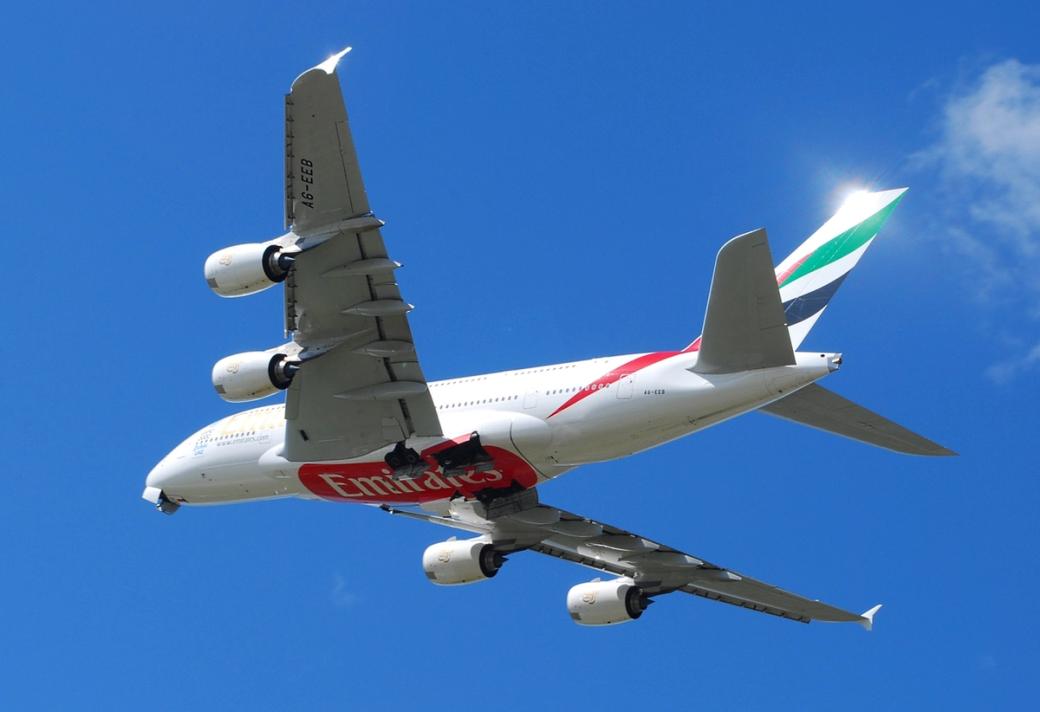 Emirates Airbus A380 jet in flight against a clear blue sky, seen from below.