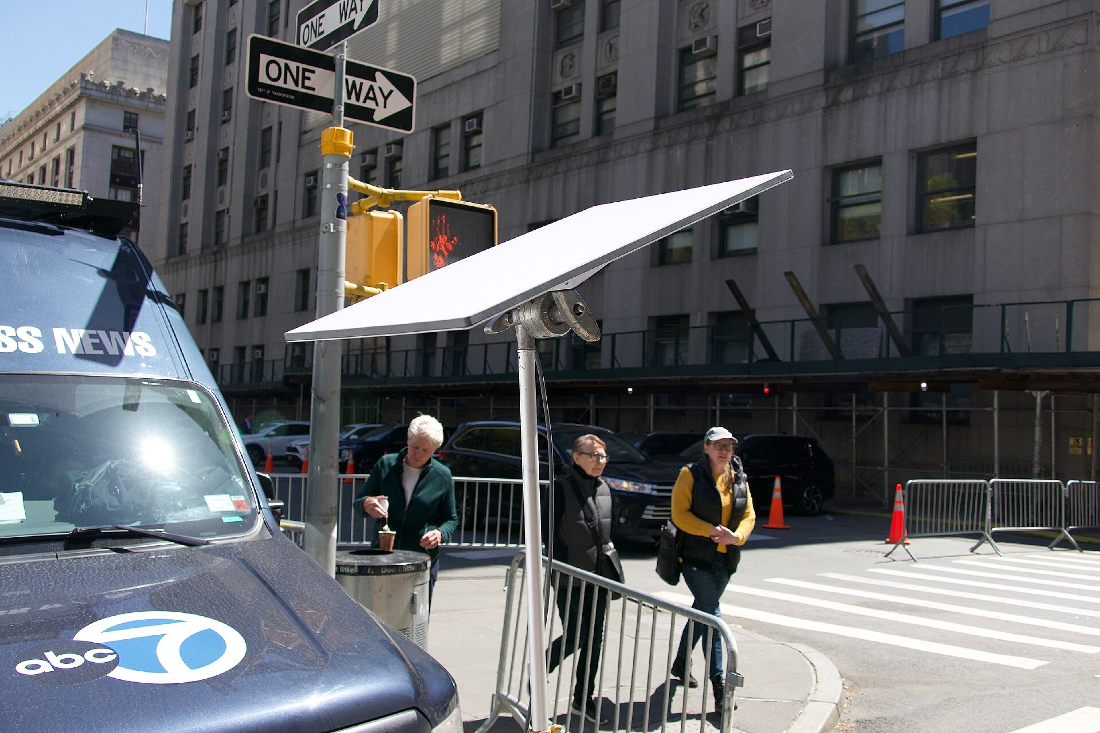 Flat satellite dish mounted on a pole beside a city street, with a news van and pedestrians nearby