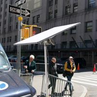 Flat satellite dish mounted on a pole beside a city street, with a news van and pedestrians nearby