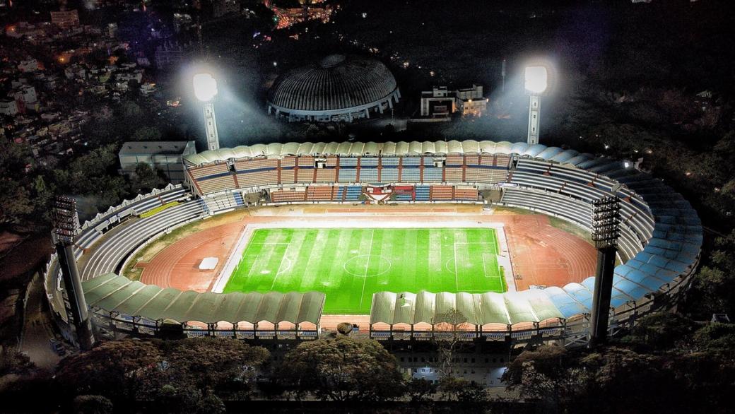 Aerial night view of a floodlit football stadium with a green pitch, running track and tiered stands.