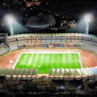 Aerial night view of a floodlit football stadium with a green pitch, running track and tiered stands.