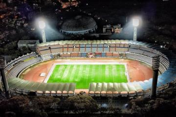 Aerial night view of a floodlit football stadium with a green pitch, running track and tiered stands.