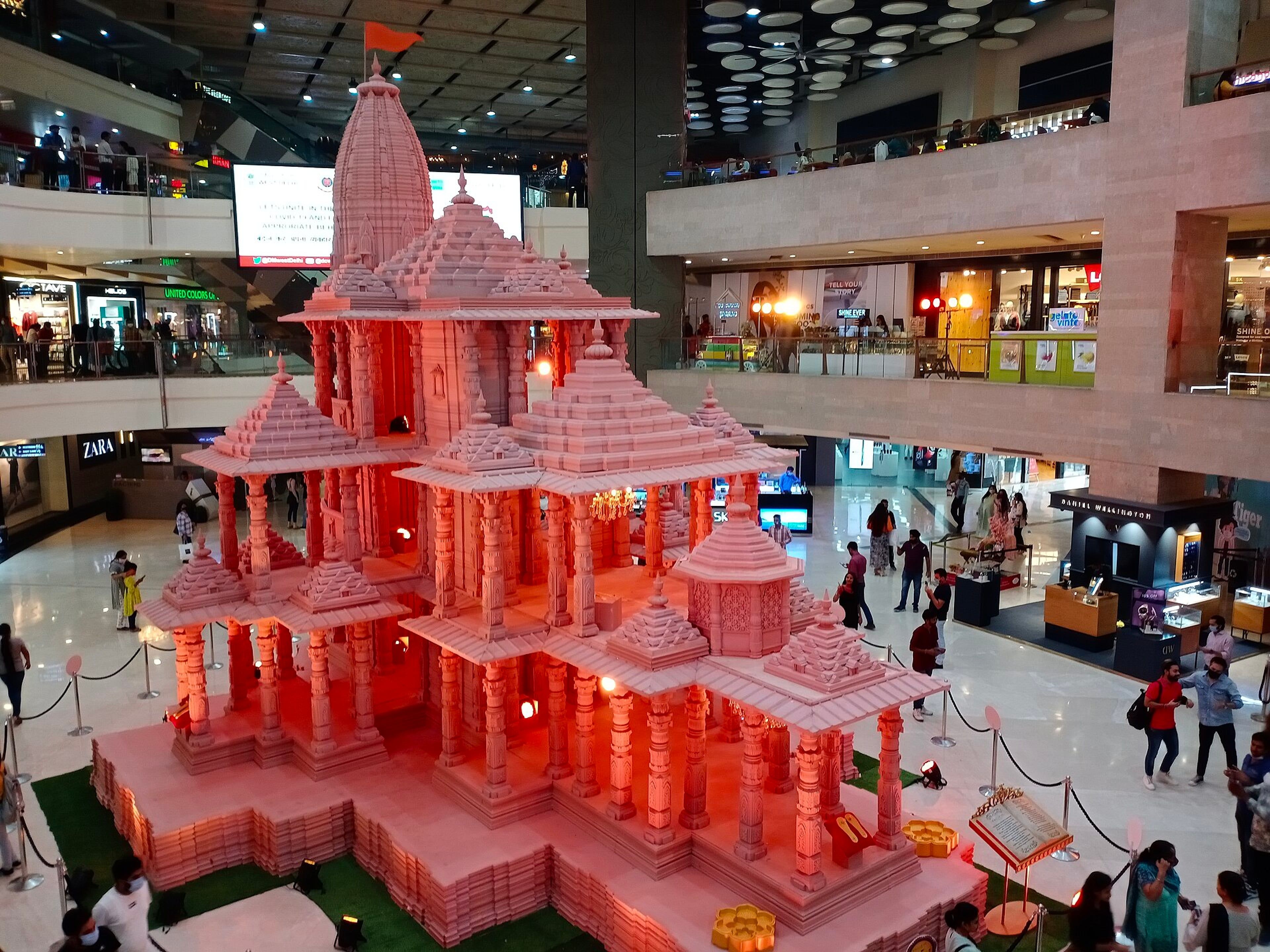 Miniature Ram Temple model on display in a New Delhi shopping mall, with shoppers on the floor and upper levels.
