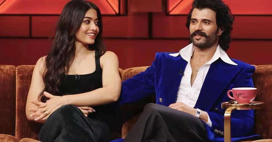 Rashmika Mandanna and Vijay Deverakonda seated on a sofa on a red-lit talk show set, with a pink coffee cup on a side table