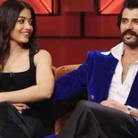 Rashmika Mandanna and Vijay Deverakonda seated on a sofa on a red-lit talk show set, with a pink coffee cup on a side table